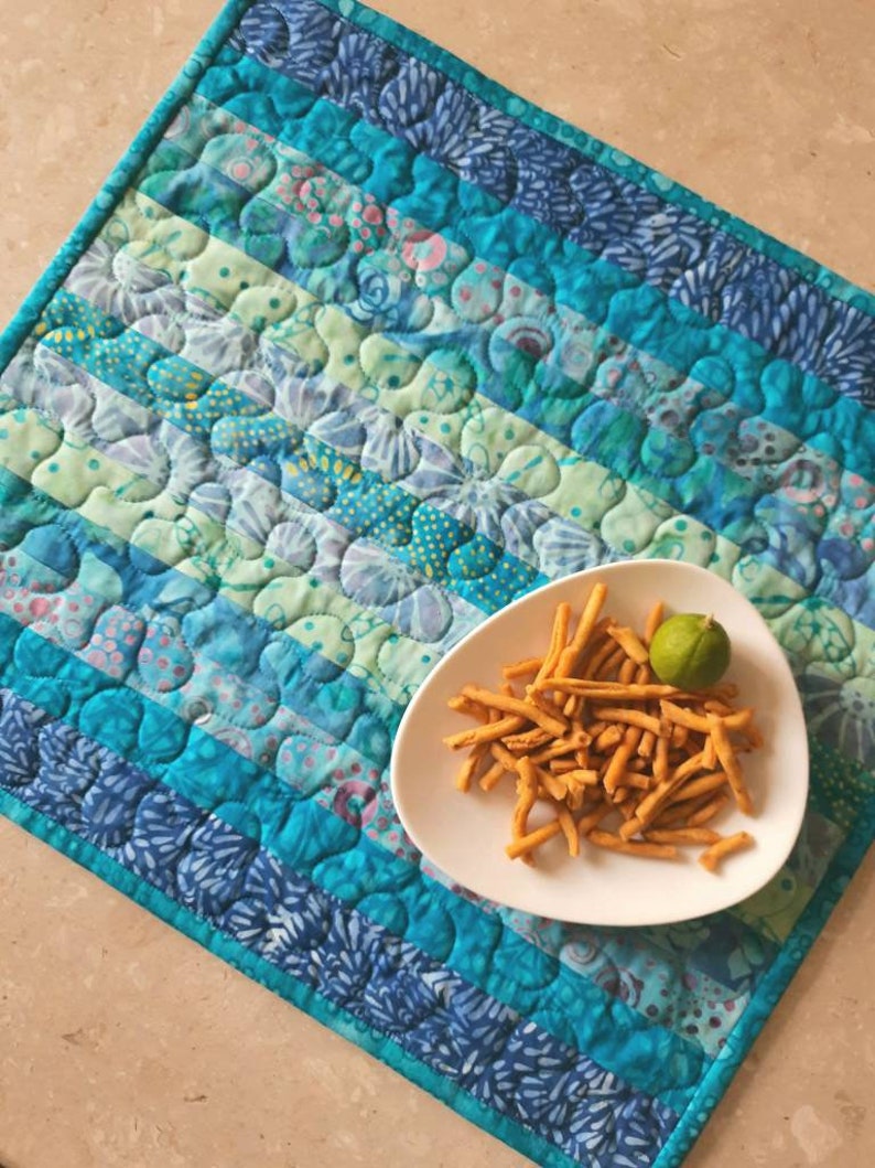 May include: A blue and green quilted table runner with a white plate of fried snacks and a lime wedge on top.