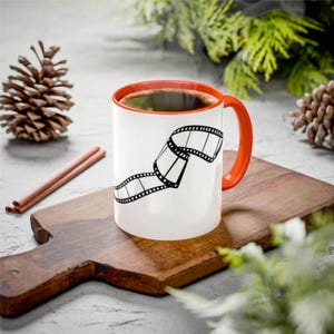 May include: A white coffee mug with an orange rim and handle. The mug has a black and white illustration of a film strip on it. The mug is sitting on a wooden cutting board.