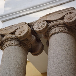Op de afbeelding: Twee stenen kolommen met sierlijke kapitelen, met ingewikkelde houtsnijwerk en krulwerk. De kolommen zijn verweerd en hebben een rustieke uitstraling.