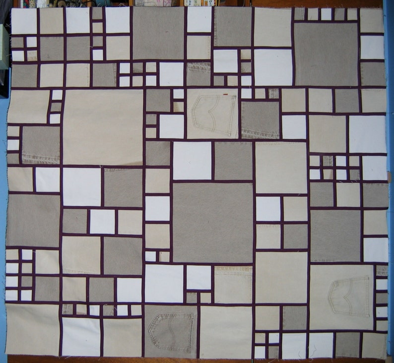 May include: A quilt made of various shades of beige fabric with a patchwork design. The quilt is made up of squares and rectangles of different sizes, and the seams are outlined in a dark purple color. Some of the squares are made of denim fabric with a pocket detail.