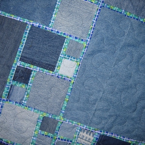 May include: A blue denim quilt with a patchwork design. The quilt is made of different shades of blue denim and is quilted with a wavy pattern.