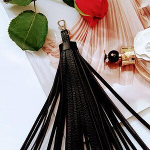 May include: A black leather tassel with a gold-coloured clasp. The tassel is made of multiple long, thin strips of leather. A red rose and green leaves are in the background, along with a perfume bottle.