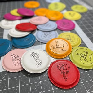 Peut inclure: Une collection de dessous de verre ronds en cuir colorés. Certains dessous de verre présentent des illustrations d'un mètre ruban, d'une rose et d'une ballerine. D'autres ont le texte "Rokdarbi Ieva Larsevica". Les couleurs incluent le rose, le rouge, le bleu et le jaune.