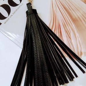 May include: A black leather tassel keyring with a silver clasp. The tassel is made of long, thin strips of textured leather. The background includes a magazine with the word "LOOK" and the text "PAVASARIS VASARA 2017".