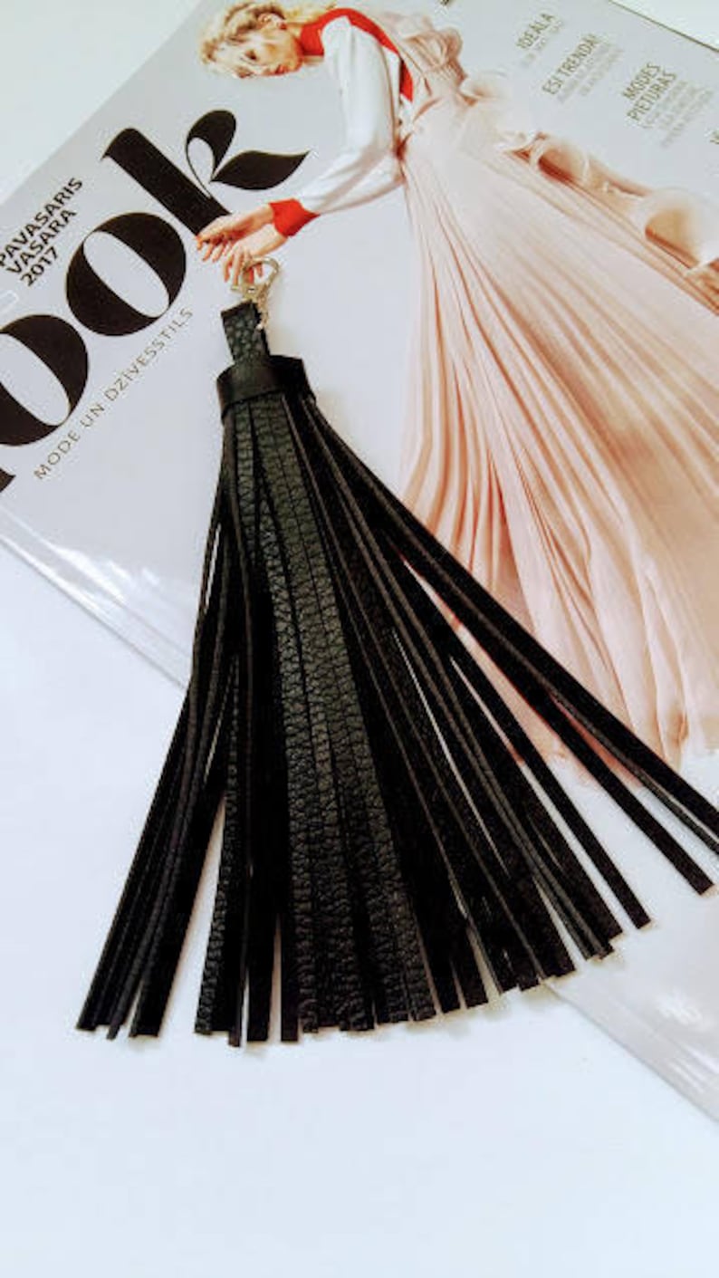 May include: A black leather tassel keyring with a silver clasp. The tassel is made of long, thin strips of textured leather. The background includes a magazine with the word "LOOK" and the text "PAVASARIS VASARA 2017".