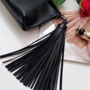 May include: A black leather phone case with a long, black tassel. The case has a textured finish and a small gold-coloured stud. A red rose and perfume bottle are in the background.