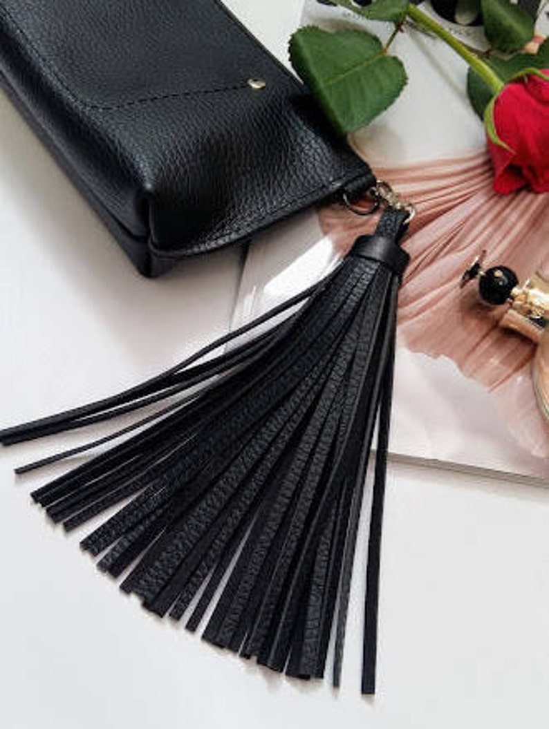 May include: A black leather phone case with a long, black tassel. The case has a textured finish and a small gold-coloured stud. A red rose and perfume bottle are in the background.