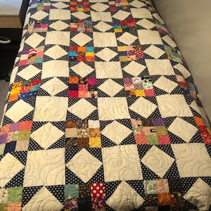 May include: A handmade quilt with a star pattern. The quilt features a cream-colored background with navy blue polka dot accents and colorful patchwork squares. The quilt is on a bed with a wooden headboard.