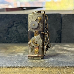 Puede incluir: Un objeto cilíndrico de estilo steampunk con engranajes, detalles metálicos y una cadena. El objeto está compuesto por varios componentes metálicos, incluyendo latón y cobre, y presenta intrincados detalles mecánicos. La estética general es envejecida e industrial.