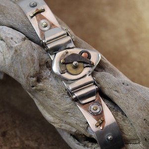 May include: A silver bracelet with a steampunk design featuring gears, cogs, and copper accents. The bracelet is on a light brown wooden surface.