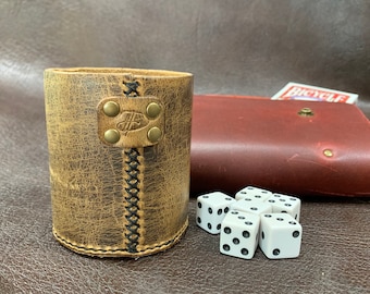 May include: A brown leather dice cup with black stitching and brass accents. Six white dice are scattered on a brown surface.