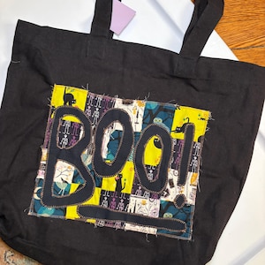 May include: A black tote bag featuring a central rectangular patch with a patchwork pattern of Halloween-themed illustrations including black cats, skeletons, and potions. The word BOO! is appliqued in black fabric over the patchwork design.