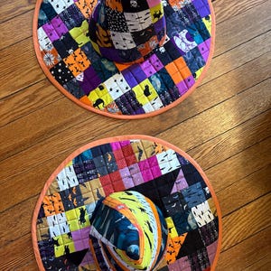 May include: Two quilted witch hats featuring a patchwork design of Halloween-themed fabric squares. The patterns include skeletons, black cats, bats, moons, and stars in vibrant shades of orange, purple, black, and white with orange trim.