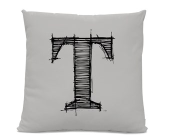 May include: A square, light gray throw pillow featuring a large, hand-drawn black letter "T" in a sketched style. The letter is centered on the pillow and has a textured appearance due to the cross-hatching.