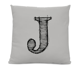 May include: A square, light gray throw pillow featuring a large, hand-drawn black letter "J". The letter is filled with horizontal lines, giving it a sketched appearance. The pillow has a soft, neutral color palette.