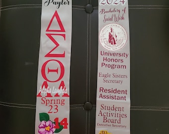 May include: A silver graduation stole with the text "Courtney Kiara Paylor" and "Alpha Lambda Spring 23" on the left side. The right side has the text "Class of 2024", "Bachelors of Social Work", "University Honors Program", "Eagle Sisters Secretary", "Resident Assistant", "Student Activities Board", "Executive Secretary", and "Miss Omega Psi Phi Fraternity, Inc."
