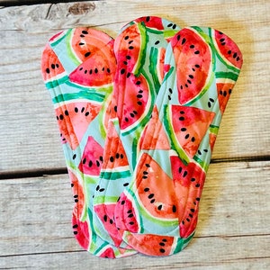 以下が含まれることがあります： スイカ柄の布製生理用ナプキン3枚。ナプキンは薄い青色で、赤と黒のスイカが描かれています。