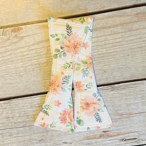 以下が含まれることがあります： ピーチ、クリーム、グリーンの色合いの花柄の蝶ネクタイ。蝶ネクタイは布製で、縁に白いステッチが施されています。