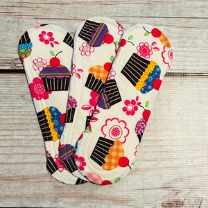 以下が含まれることがあります： カップケーキ柄の白い布製の生理用ナプキンが3枚。カップケーキは茶色、ピンク、青、黄色の白いフロスティングと赤いチェリー。背景は白地にピンクと緑の花柄です。