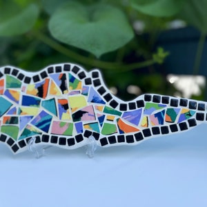 Puede incluir: Una pieza de arte de mosaico de azulejos coloridos con forma de isla con un borde negro. Los azulejos de mosaico son de varios tonos de azul, verde, amarillo, naranja, rosa y morado.