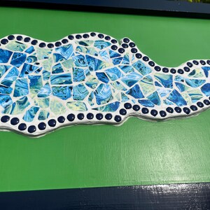 Puede incluir: Una pieza de arte mural de mosaico con la forma del estado de Kentucky, que presenta un borde de azulejos circulares azul oscuro y un interior lleno de fragmentos irregulares de vidrio veteado azul, azul claro y blanco sobre una base de lechada blanca.