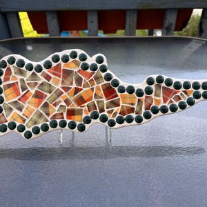 Puede incluir: Mosaico decorativo con forma de nube o isla estilizada. El mosaico presenta un diseño de patchwork de baldosas a cuadros naranjas, rojas y marrones, delineadas con acentos circulares de color verde oscuro. Montado sobre soportes transparentes.