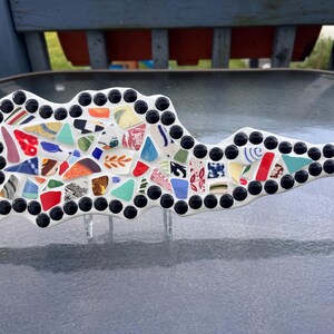 Puede incluir: Una escultura de mosaico con forma de isla, con una base blanca con piezas de cerámica de colores. El mosaico está delineado con cuentas negras y se apoya en soportes de acrílico transparente. La obra de arte se exhibe sobre una superficie reflectante.