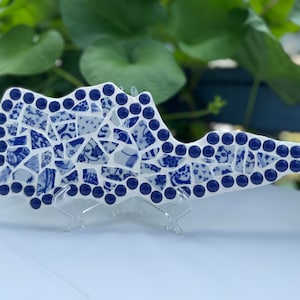 Puede incluir: Una pieza de arte decorativo con la forma del estado de Kentucky, elaborada con un mosaico de fragmentos de cerámica con patrones azules y blancos. El borde está delineado con pequeñas cuentas de vidrio azul oscuro sobre un soporte acrílico.