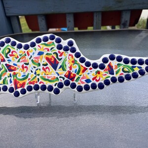 Puede incluir: Una pieza decorativa de cerámica con una forma única, con un diseño de mosaico. El mosaico está compuesto por azulejos irregulares de colores rojo, amarillo, azul y verde, delineados con un borde blanco y puntos azul oscuro. Se apoya en soportes transparentes.