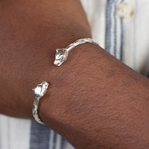 May include: Silver bracelet with two panther heads on the ends. The bracelet is open and has a textured design.