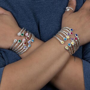 May include: Two silver-toned cuff bracelets featuring multiple rows of metal bands, each tipped with a small, round, multi-colored gemstone. The bracelets are worn on crossed wrists, and a silver ring with a floral design is visible on one finger.