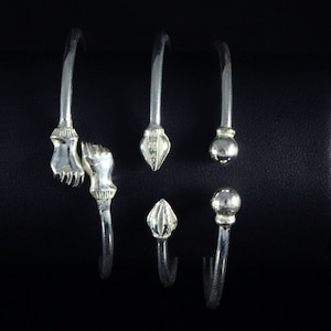 May include: Three silver bangles with different decorative ends. One bangle has two hands clasped together, another has a pointed end, and the third has a round ball end.