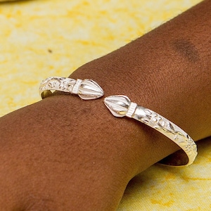 May include: A silver bangle bracelet with a floral design and two decorative ends. The bracelet is worn on a person's wrist.