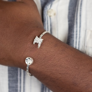 May include: A silver bangle bracelet with a textured design in the shape of an island. The bracelet is open and has a circular clasp.