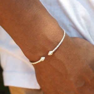 May include: A silver-toned open cuff bracelet featuring a textured, cross-hatched pattern along the band and two pointed, bud-shaped terminals at each end. The metal surface reflects light, highlighting the intricate detailing of the design.