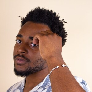 May include: A man with short, curly black hair is wearing a silver cuff bracelet on his left wrist. He is wearing a light blue and white striped shirt. His hand is raised to his forehead, and he is looking off to the side.