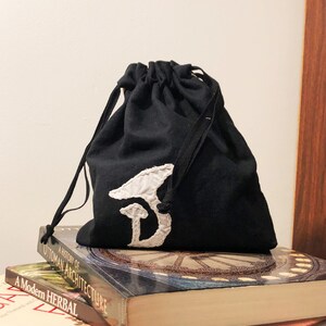 Pode incluir: Uma bolsa de cordão preta com uma letra D bordada em branco na frente. A bolsa está em cima de uma pilha de livros.