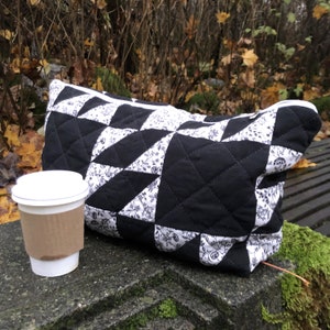 May include: A black and white quilted zippered pouch with a floral pattern sits on a stone surface next to a disposable coffee cup.