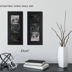 May include: Two black framed wall art pieces with geometric spiral designs in silver and white. The text "3D GEOMETRIC LONG WALL ART" is above the art, and the word "Duo" is below. A white vase with branches and decorative spheres are also present.