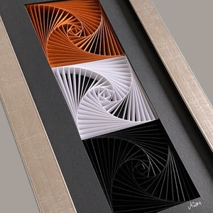 May include: A framed artwork featuring three concentric spirals in orange, white, and black. The spirals are created using paper strips and are arranged in a layered effect.