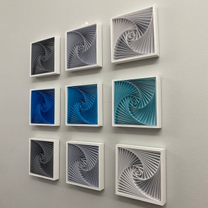 Peut inclure: Neuf œuvres d'art encadrées en papier avec un motif en spirale dans des tons de gris, de bleu et de sarcelle. Chaque pièce est carrée et encadrée dans un cadre blanc. L'œuvre est accrochée à un mur gris clair.