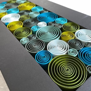 May include: A framed wall art piece featuring a variety of paper rolls in shades of green, blue, and gray. The rolls are arranged in a circular pattern, creating a visually appealing and textured design.