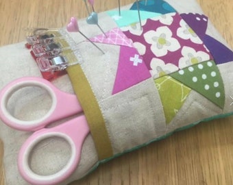 May include: A beige fabric pincushion with a patchwork design featuring various colors and patterns, including pink, green, purple, and white. The pincushion has a pocket for scissors and a clear plastic clip holder.  There are several pins stuck into the pincushion.