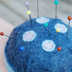 May include: A blue felt pincushion with white felt circles and colorful pins. The pincushion is a sphere shape and has a white felt trim around the bottom.