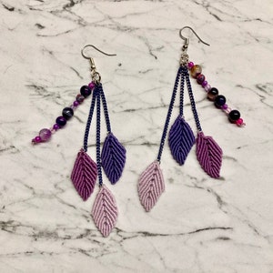 Peut inclure: Une paire de boucles d'oreilles longues et pendantes avec plusieurs feuilles violettes et roses et des perles. Les boucles d'oreilles sont faites avec un crochet argenté et une chaîne de perles violettes et roses.
