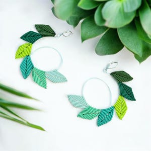 May include: A pair of green and blue leaf-shaped hoop earrings. The earrings are made of a woven material and have a silver hoop.