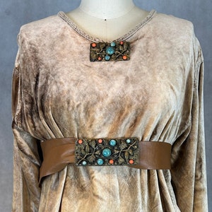 Könnte beinhalten: Eine beige Samt-Tunika mit langen Ärmeln und V-Ausschnitt. Die Tunika hat einen braunen Ledergürtel mit einer dekorativen Metallschnalle mit türkisfarbenen und orangefarbenen Steinen.