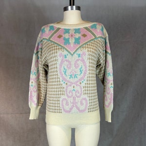 May include: Cream-colored sweater with a boat neck and long sleeves. The sweater features a houndstooth pattern with pink, teal, and gold geometric and floral designs. The cuffs and neckline are solid cream.
