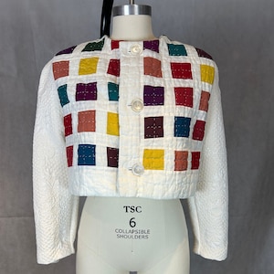 May include: A white quilted jacket with a patchwork design featuring squares of various colors, including red, green, yellow, purple, and brown. The jacket has a button closure and a cropped length. The label on the jacket reads "TSC 6 COLLAPSIBLE SHOULDERS".