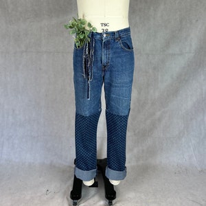 May include: Blue denim jeans with a patchwork design on the lower legs. The jeans have a frayed hem and a small green plant tucked into the front pocket.
